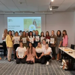 Grupa kobiet, prawdopodobnie lektorek, pozuje z certyfikatami ukończenia kursu metody Teddy Eddie na tle wyświetlanego logo Edu Bears oraz prezentacji multimedialnej.