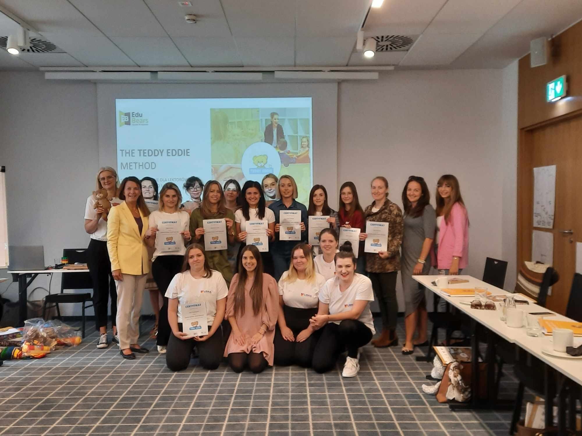 Grupa kobiet, prawdopodobnie lektorek, pozuje z certyfikatami ukończenia kursu metody Teddy Eddie na tle wyświetlanego logo Edu Bears oraz prezentacji multimedialnej.