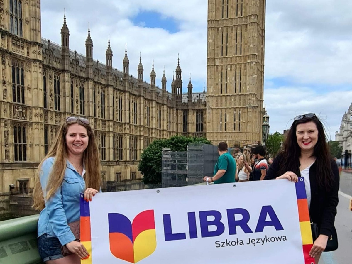 Dwie uśmiechnięte kobiety trzymają baner szkoły językowej Libra na tle Big Bena i budynków Parlamentu w Londynie.