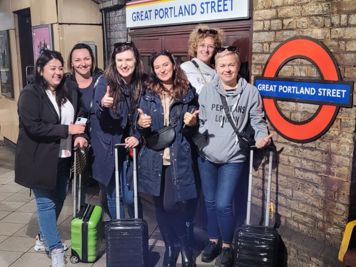 Grupa sześciu uśmiechniętych kobiet z walizkami pozuje na tle znaku stacji metra Great Portland Street, pokazując kciuki w górę.