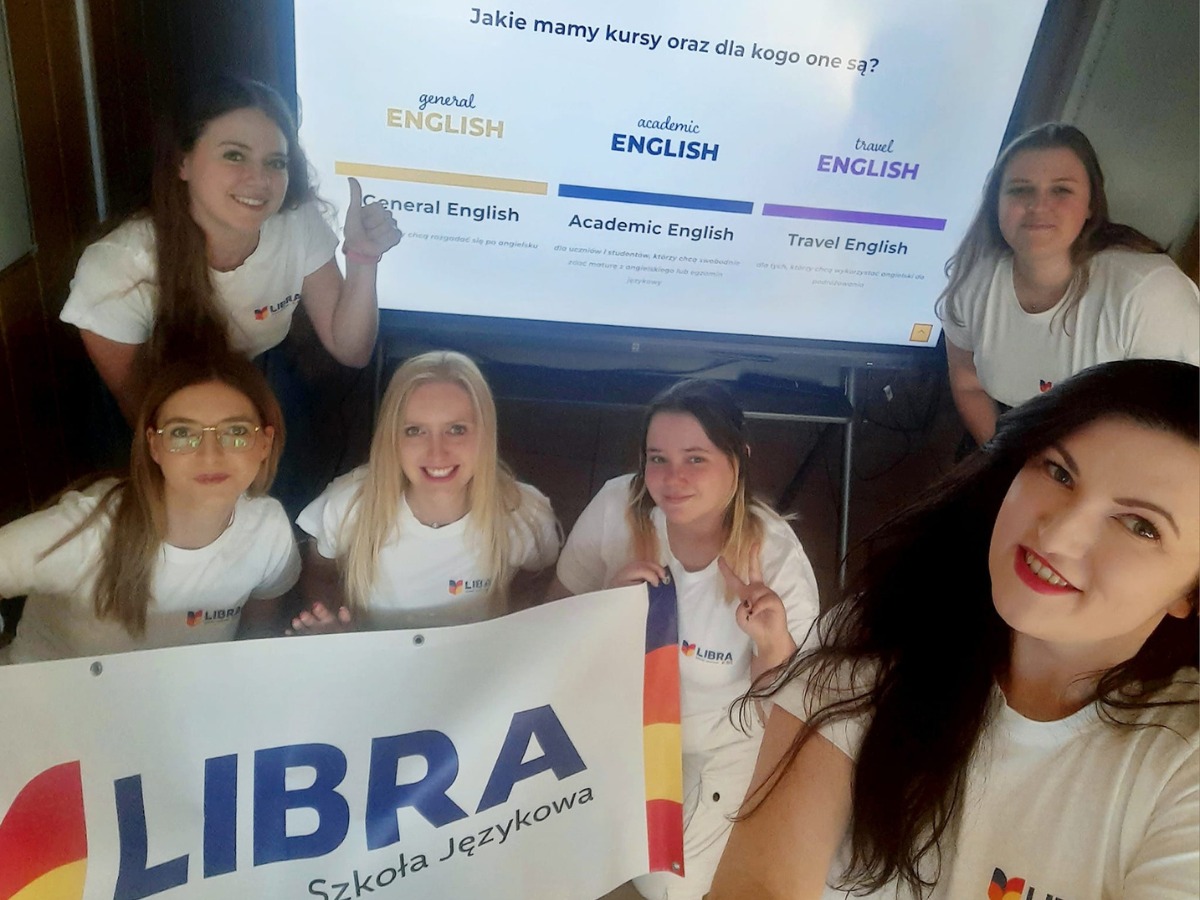Grupa uśmiechniętych kobiet w białych koszulkach z logo szkoły językowej Libra, prezentujących baner z nazwą szkoły na tle ekranu z ofertą kursów: General English, Academic English, Travel English.