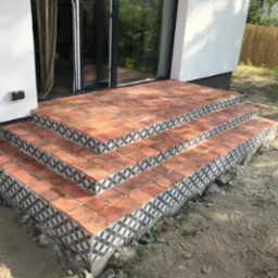 Trzy stopnie schodów wyłożone czerwoną terakotą z dekoracyjnym obramowaniem w geometryczny wzór, prowadzące do nowoczesnego domu z dużym oknem przesuwnym.