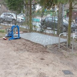 Angular Bruk - usługi brukarskie - Niewielki plac wyłożony szarą kostką brukową, obok stoi niebieska zagęszczarka, w tle drzewa i ogrodzenie z siatki.