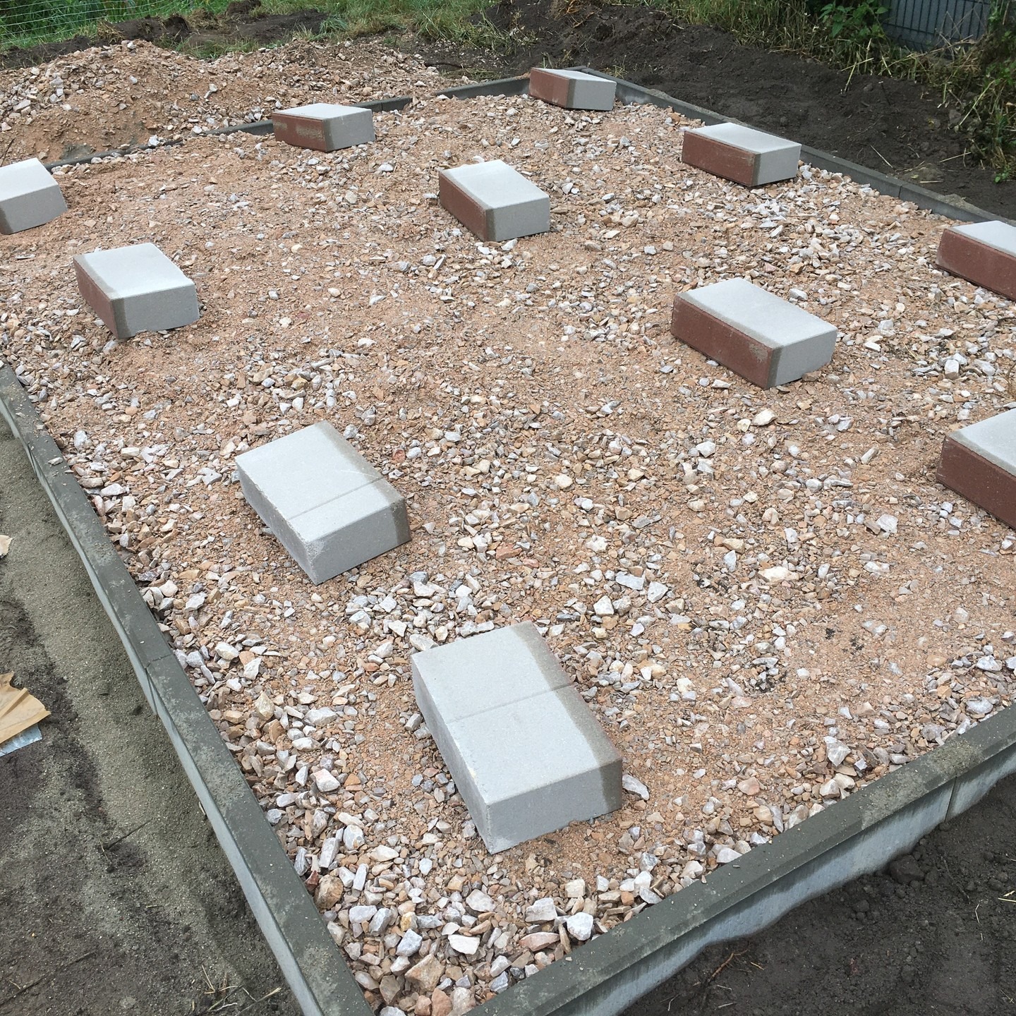 Przygotowanie podłoża pod nawierzchnię: ubity piasek z drobnymi kamieniami, obrzeża betonowe i częściowo ułożone bloczki betonowe z czerwoną cegłą na wierzchu, tworzące wzór. Widok z góry.