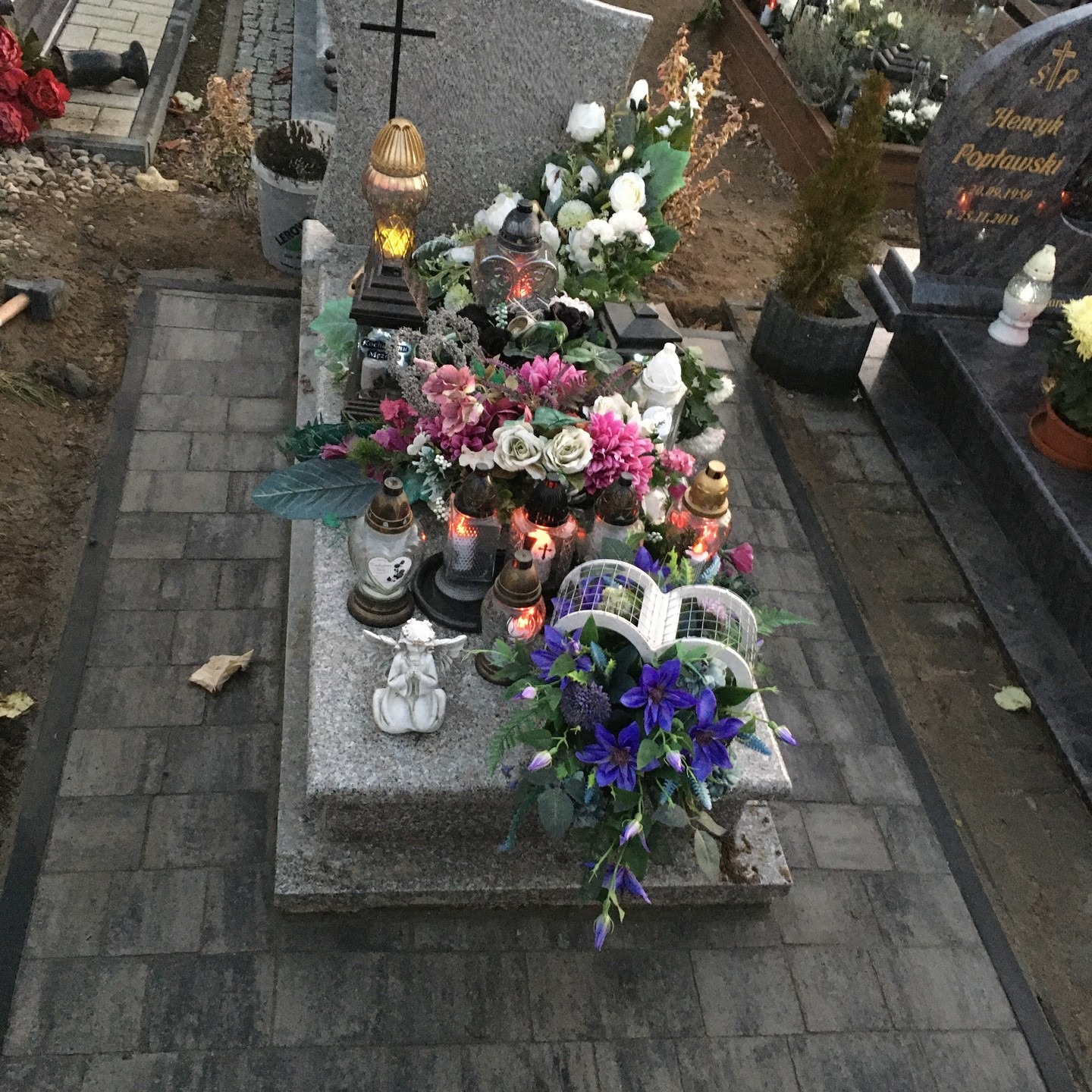 Granitowy nagrobek z zapalonymi zniczami, kwiatami i figurką anioła, otoczony nowo ułożoną szarą kostką brukową.