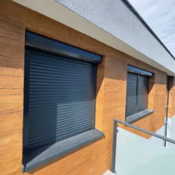 Zewnętrzne, grafitowe rolety na oknach budynku z elewacją imitującą drewno. Widok z balkonu z szklaną balustradą, częściowo zasłonięte rolety.