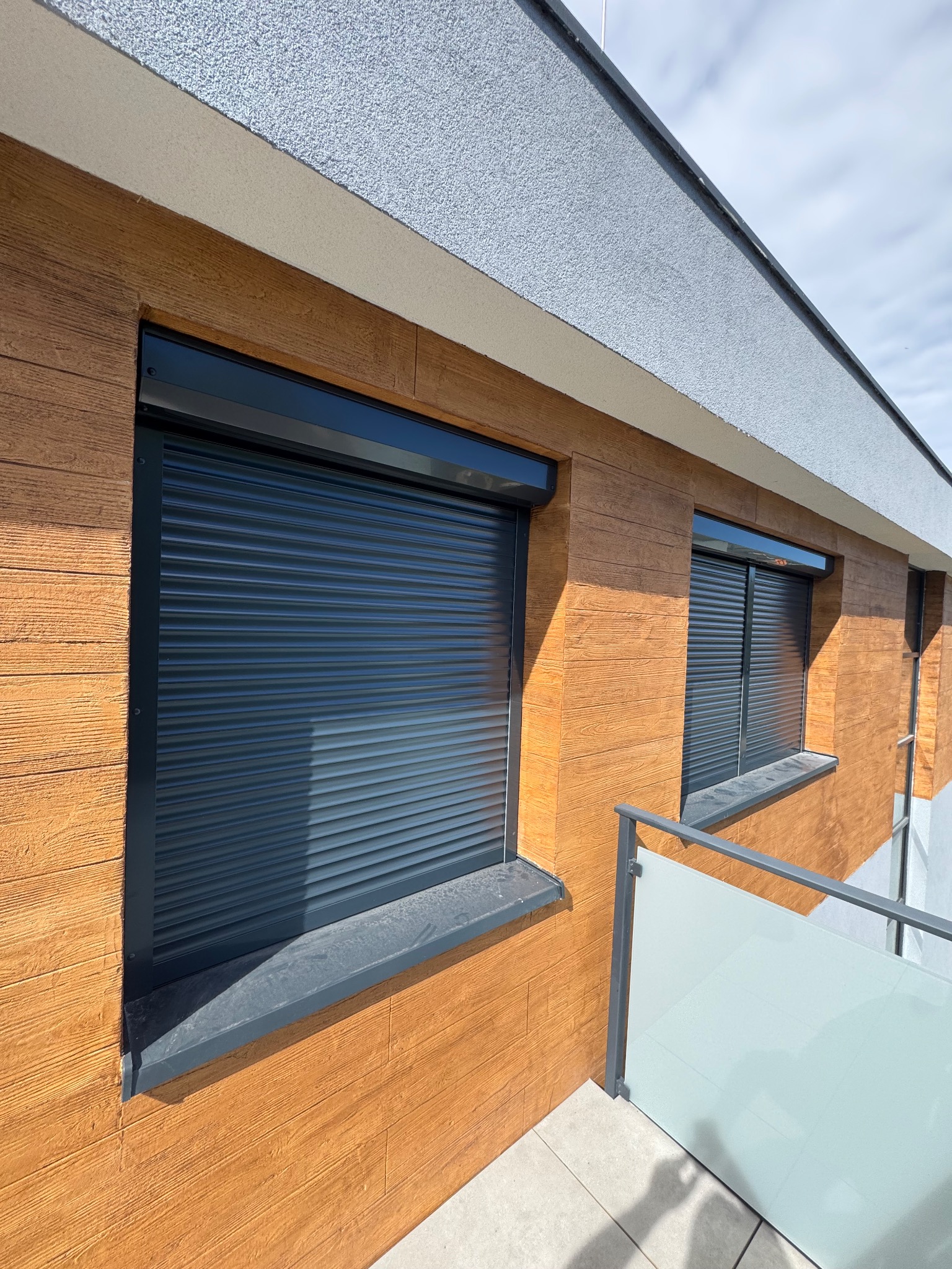 Zewnętrzne, grafitowe rolety na oknach budynku z elewacją imitującą drewno. Widok z balkonu z szklaną balustradą, częściowo zasłonięte rolety.