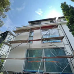 Elewacja budynku w trakcie tynkowania, okna zabezpieczone folią, widoczne rusztowania, błękitne niebo w tle.