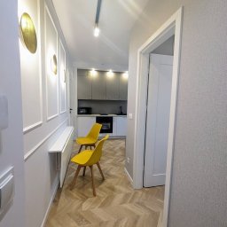 OnyxArt Piotr Padowski - Wnętrze mieszkania po remoncie: korytarz z parkietem w jodełkę, żółte krzesła i widok na szarą kuchnię z białymi blatami.