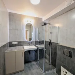 Nowoczesna łazienka z szarymi płytkami, kabiną prysznicową ze szkła, wiszącą umywalką z czarną armaturą i podświetlanym lustrem w kształcie owalu.