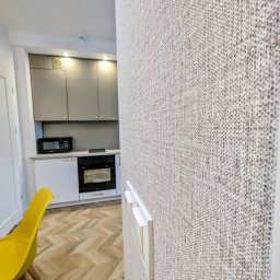 OnyxArt Piotr Padowski - Beżowa tapeta o wyraźnej fakturze na ścianie, widoczne włączniki światła, w tle fragment kuchni z szarymi szafkami i żółte krzesło na parkiecie w jodełkę.