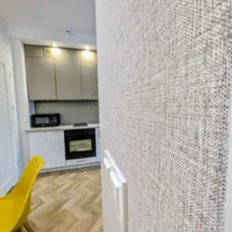 Beżowa tapeta o wyraźnej fakturze na ścianie, widoczne włączniki światła, w tle fragment kuchni z szarymi szafkami i żółte krzesło na parkiecie w jodełkę.