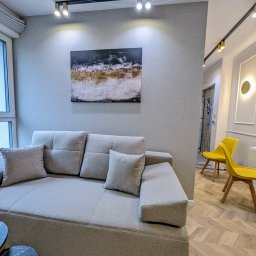OnyxArt Piotr Padowski - Jasnoszara sofa z poduszkami w salonie z szarą tapetą, abstrakcyjnym obrazem na ścianie i żółtymi krzesłami w tle, oświetlonymi złotymi lampami.