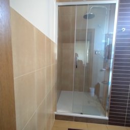 OnyxArt Piotr Padowski - Wykończona kabina prysznicowa z przesuwnymi drzwiami, beżowe płytki ścienne i brązowe płytki na podłodze, widoczny grzejnik drabinkowy wewnątrz kabiny.