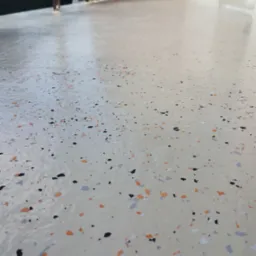 Jasna, gładka posadzka z dekoracyjnymi, kolorowymi płatkami (czarne, pomarańczowe, szare) na balkonie, widok z niskiej perspektywy, ukazujący detale wykończenia.