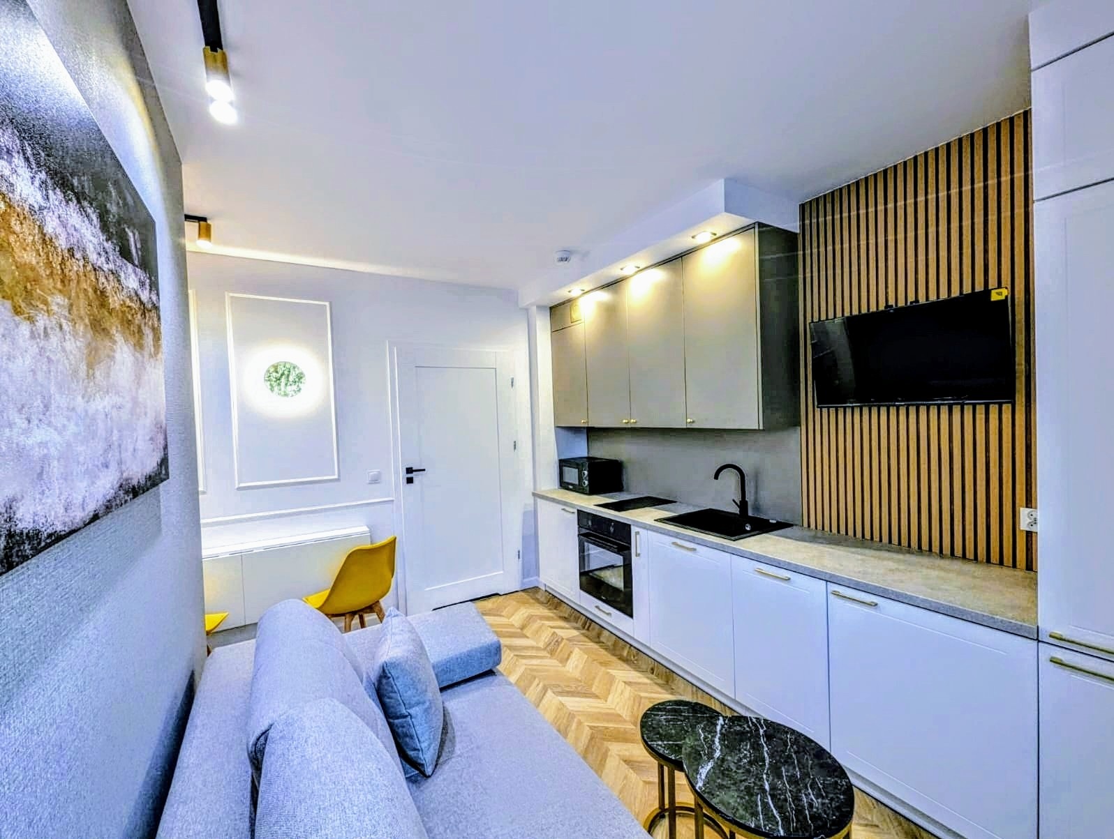 Nowoczesne wnętrze kawalerki z aneksem kuchennym: białe szafki, czarny zlew i armatura, drewniana okładzina ściany za telewizorem, podłoga w jodełkę, szara sofa i marmurowe stoliki kawowe.