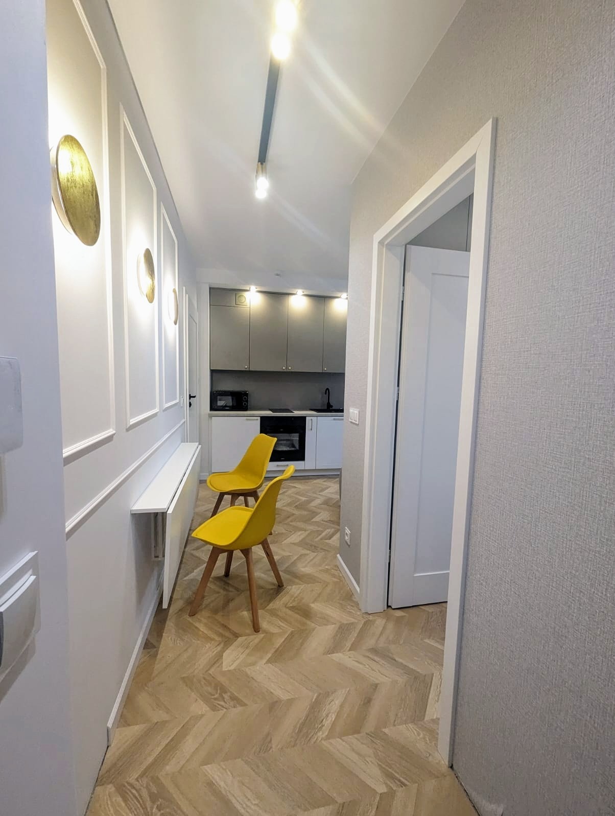 Wnętrze mieszkania po remoncie: korytarz z parkietem w jodełkę, żółte krzesła i widok na szarą kuchnię z białymi blatami.