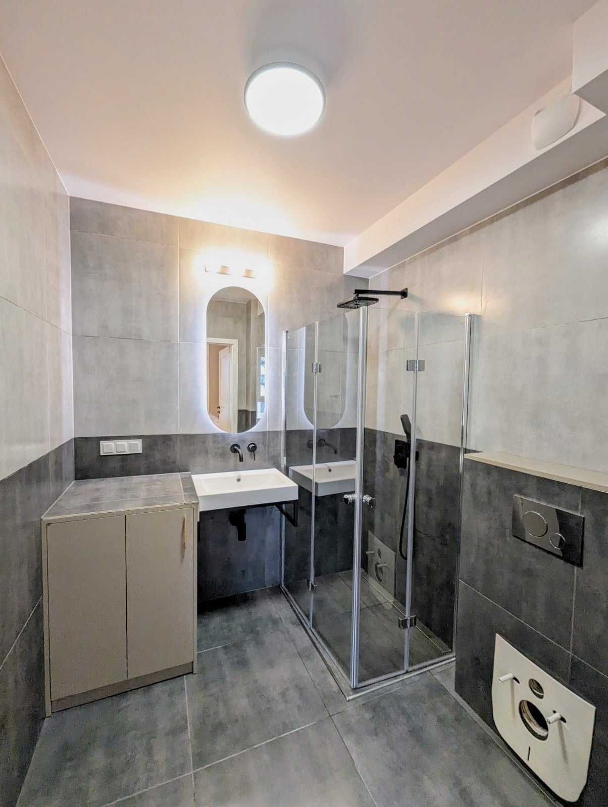 Nowoczesna łazienka z szarymi płytkami, kabiną prysznicową ze szkła, wiszącą umywalką z czarną armaturą i podświetlanym lustrem w kształcie owalu.