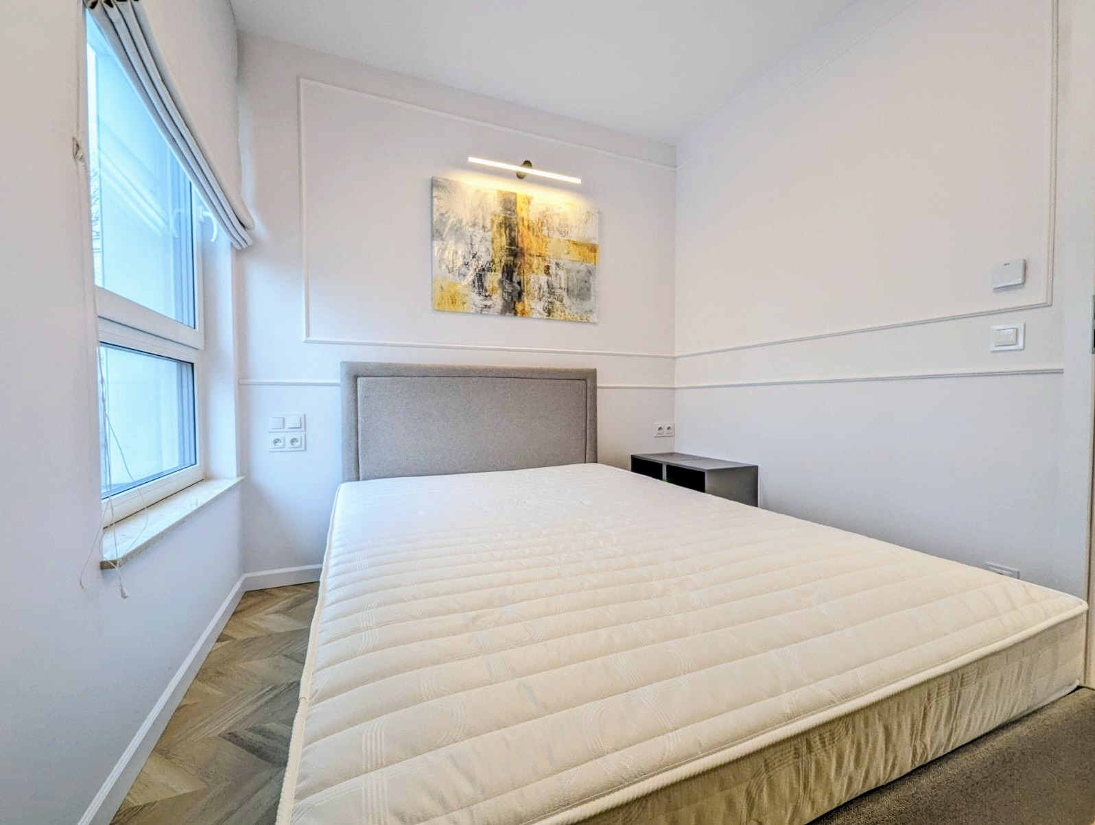 Nowoczesna sypialnia z łóżkiem kontynentalnym, szarym zagłówkiem, abstrakcyjnym obrazem w złoto-czarnych tonacjach i parkietem ułożonym we wzór jodełki.