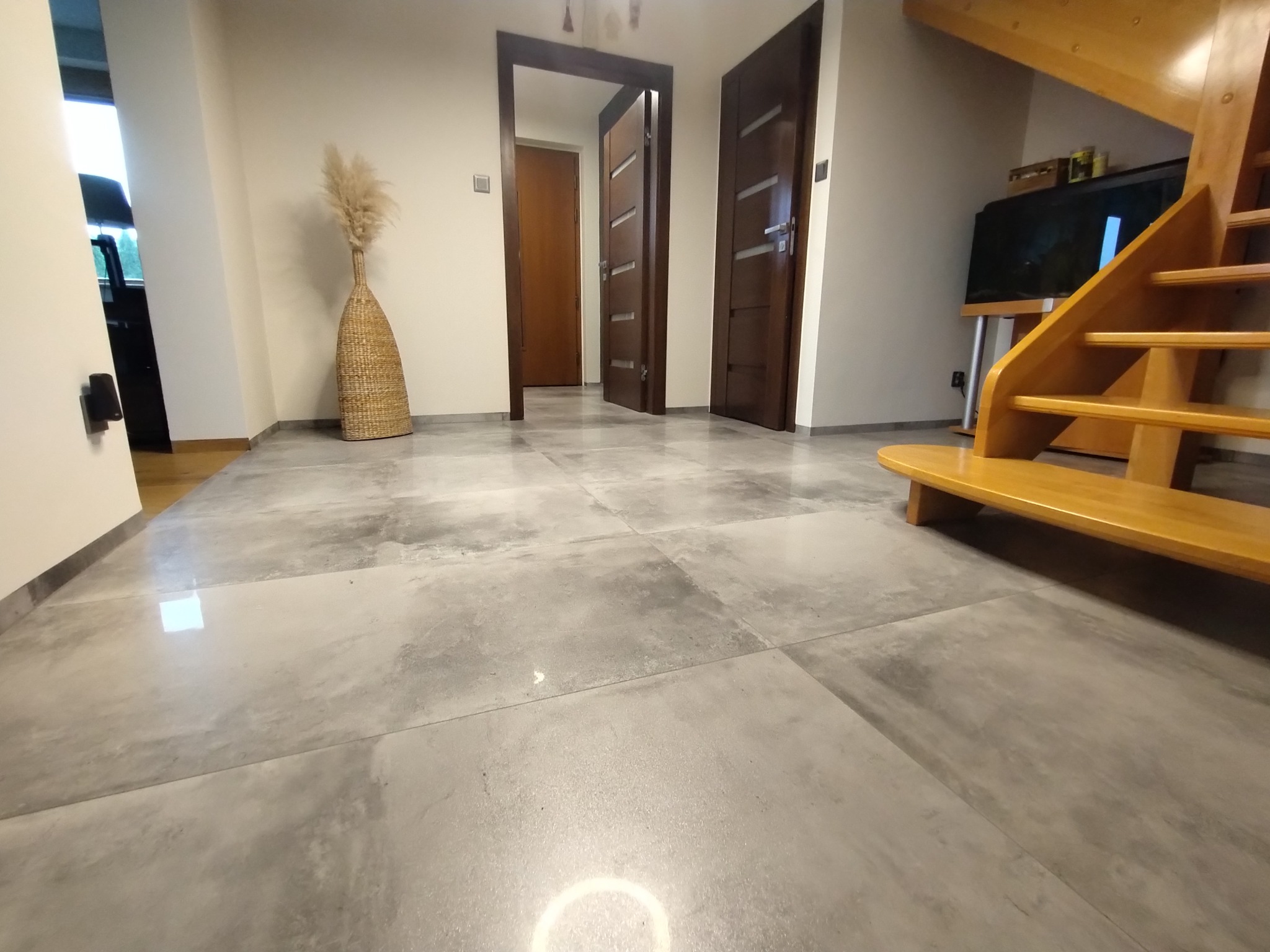 Szara, duża płytka podłogowa w przedpokoju z drewnianymi schodami, widoczne drzwi wewnętrzne i dekoracyjny wazon.