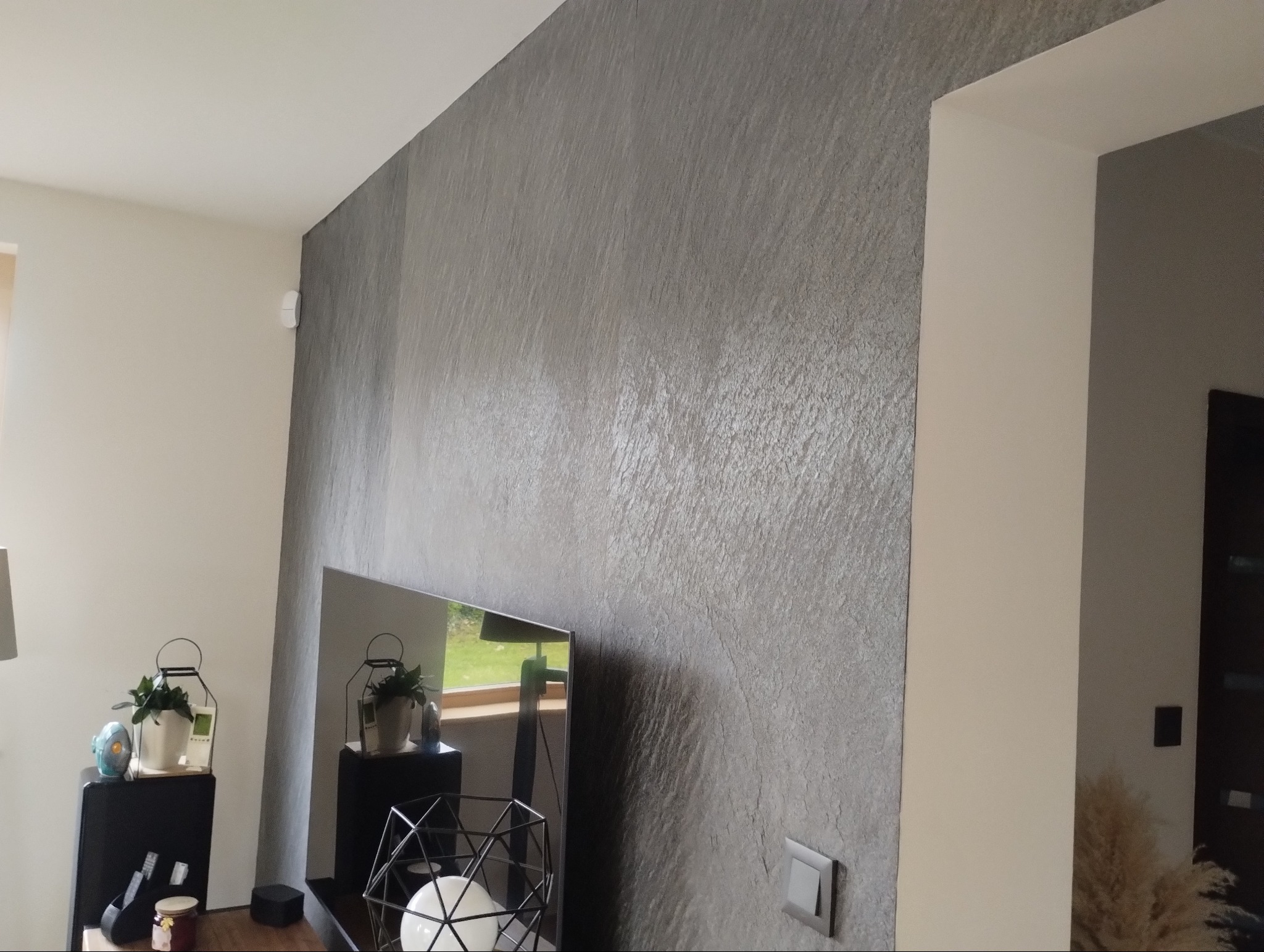 Ściana w salonie w Koleczkowie pokryta dekoracyjną szarą gładzią imitującą kamień, widoczny włącznik światła i fragment telewizora odbijający okno.