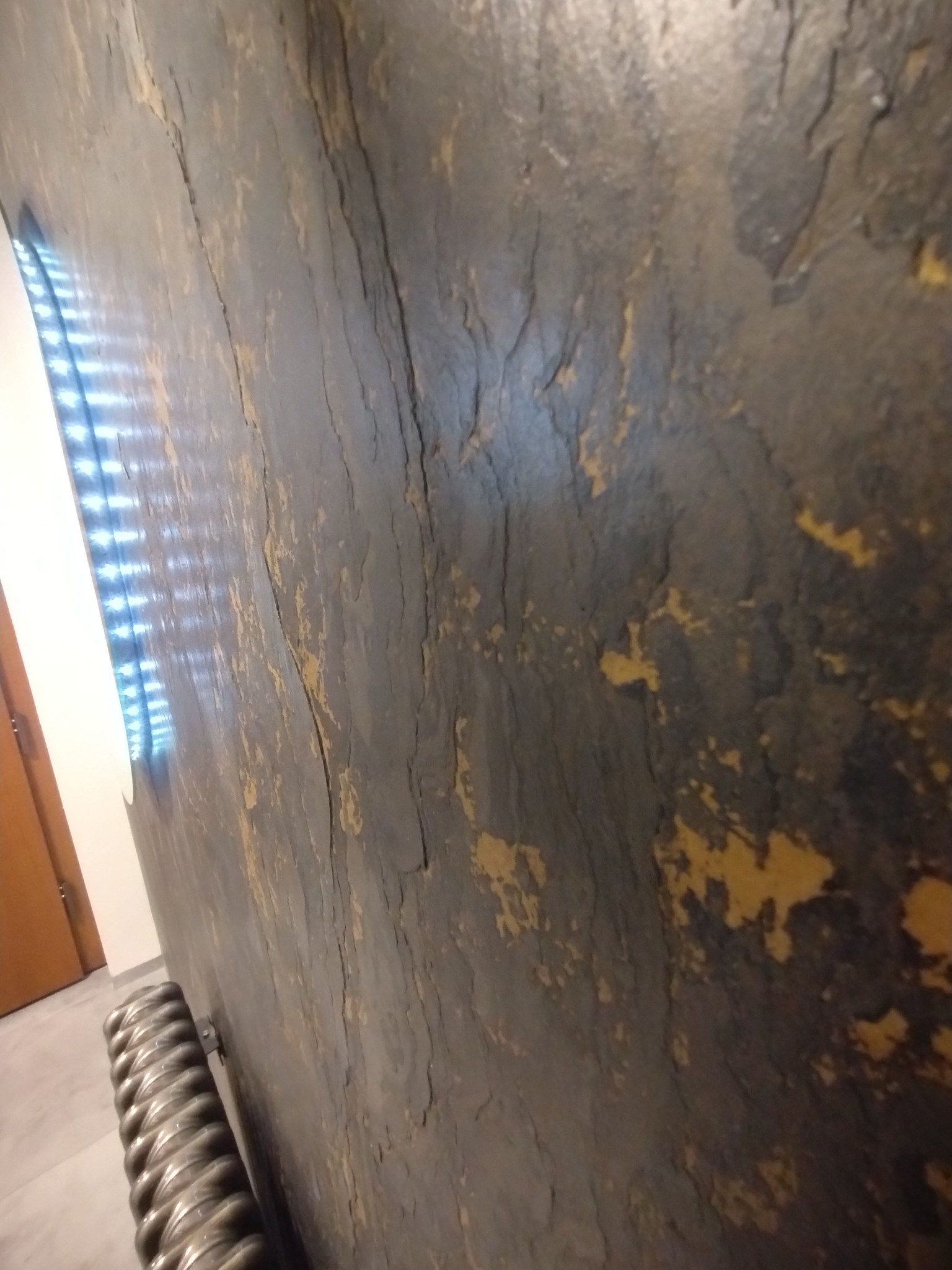 Dekoracyjna ściana w ciemnych odcieniach z wyraźną fakturą przypominającą łuskany kamień, z fragmentami złocistych przebarwień, widoczny grzejnik i fragment okna z żaluzjami.