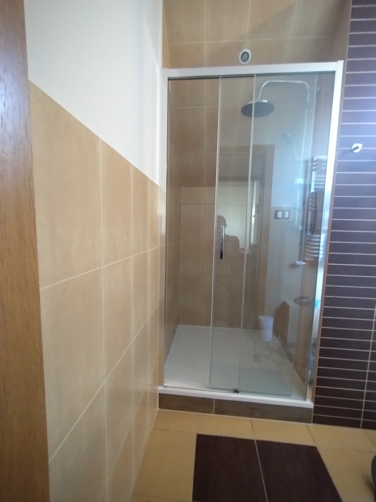 Wykończona kabina prysznicowa z przesuwnymi drzwiami, beżowe płytki ścienne i brązowe płytki na podłodze, widoczny grzejnik drabinkowy wewnątrz kabiny.