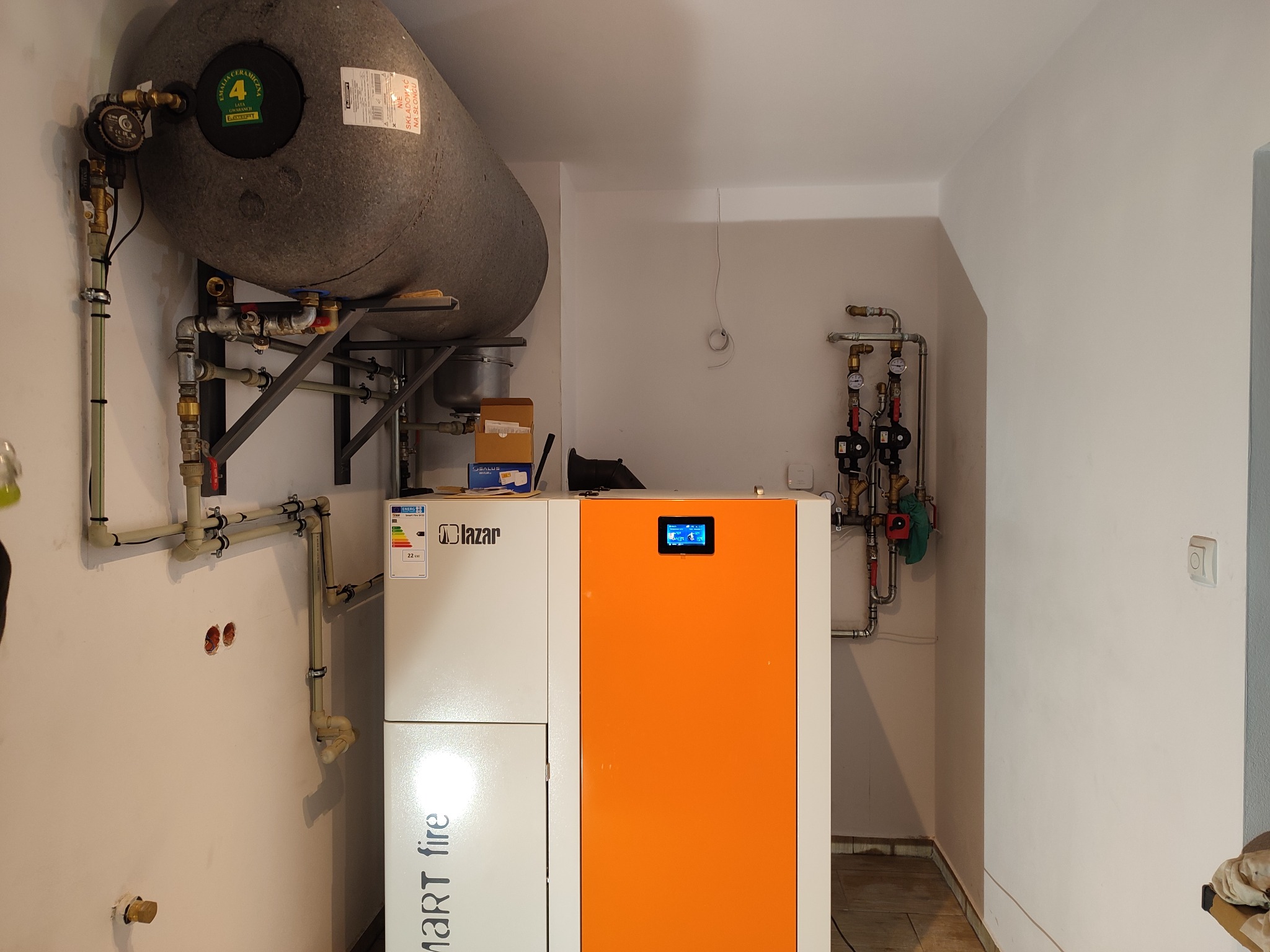 Nowoczesny piec CO marki Lazar z pomarańczowym panelem, zbiornik buforowy i instalacja grzewcza w kotłowni. Widoczne rury i zawory.