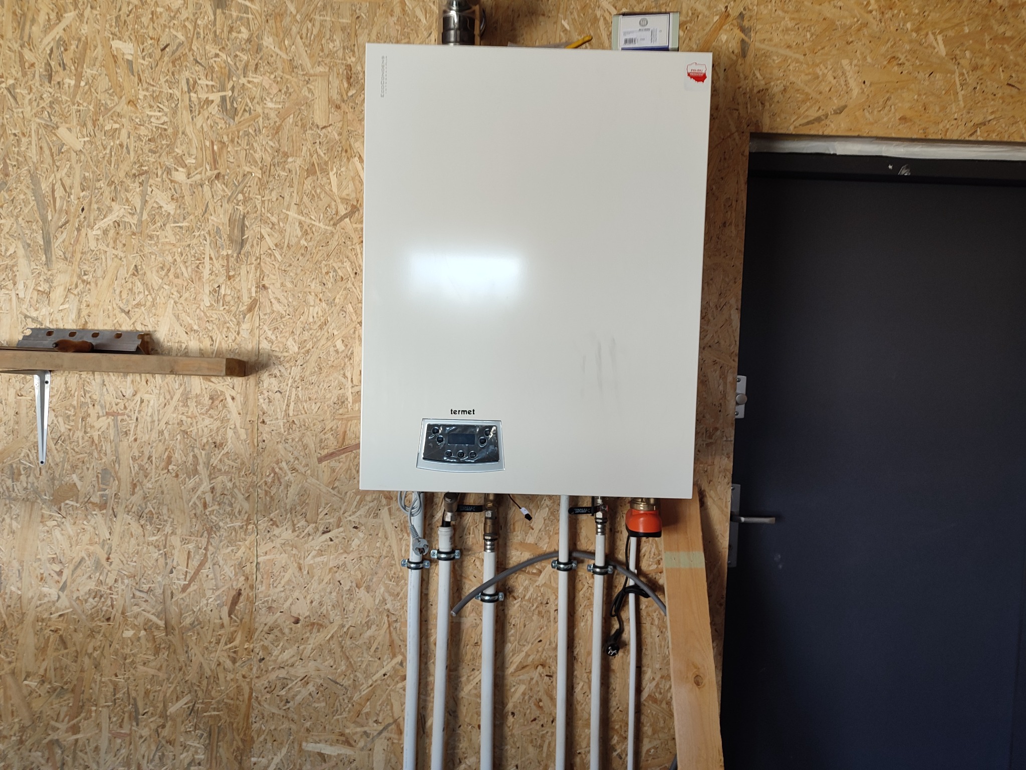 Biały piec gazowy Termet zamontowany na ścianie z płyty OSB, z widocznymi rurami instalacji. Obok ciemne drzwi. Ujęcie z bliska, prezentujące szczegóły instalacji.