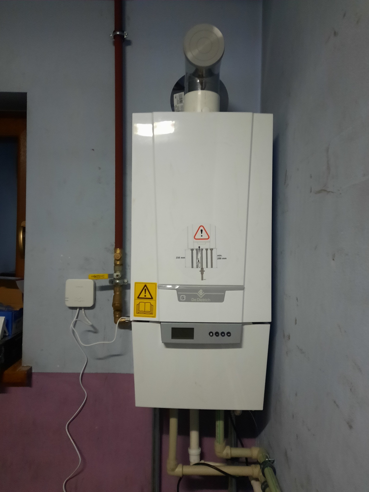 Biały piec gazowy De Dietrich zamontowany na ścianie z widocznymi rurami i elementami instalacji, oraz czujnikiem temperatury.