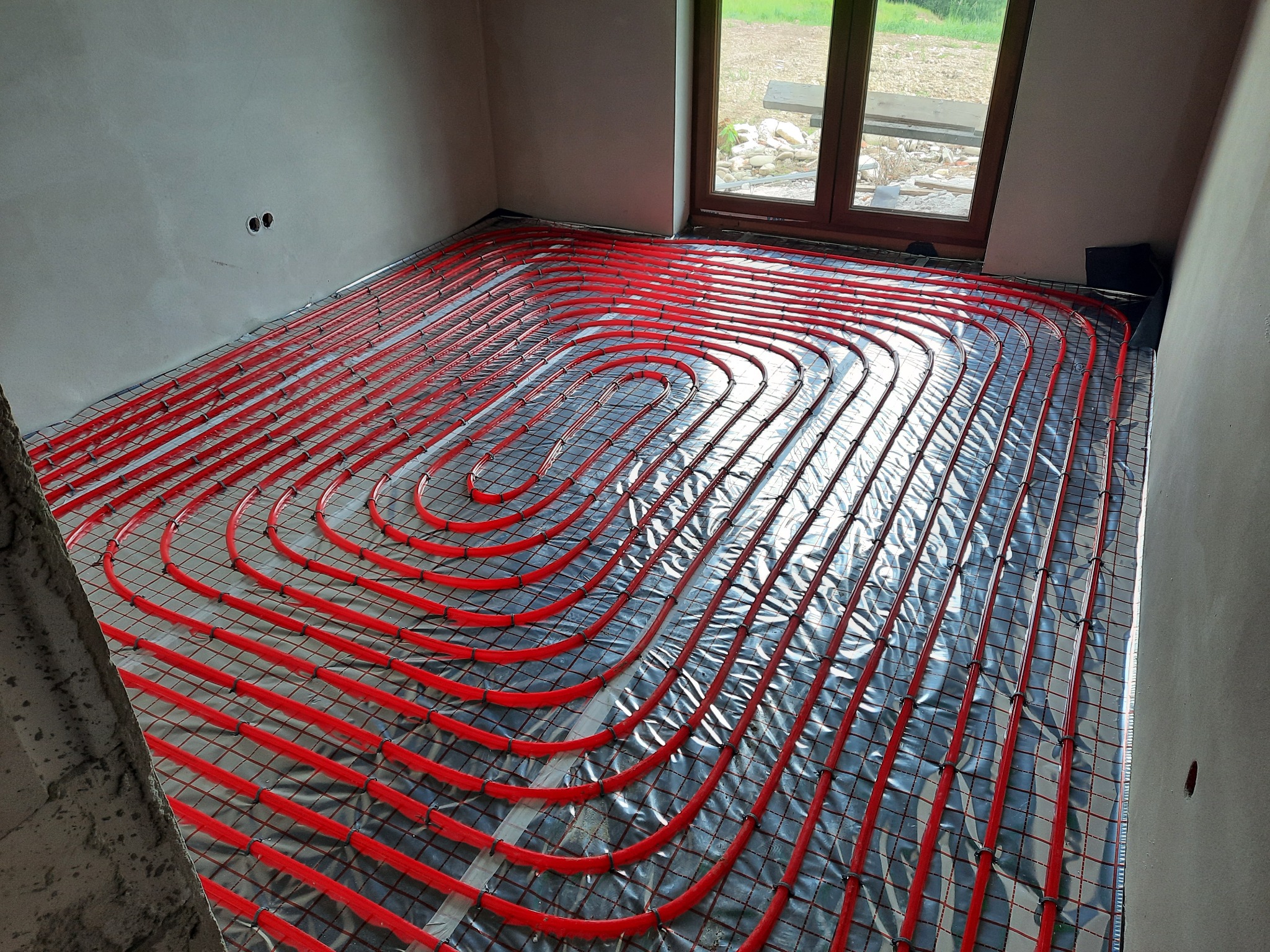 Czerwone rury ogrzewania podłogowego ułożone spiralnie na srebrnej folii izolacyjnej w surowym pomieszczeniu z oknem.