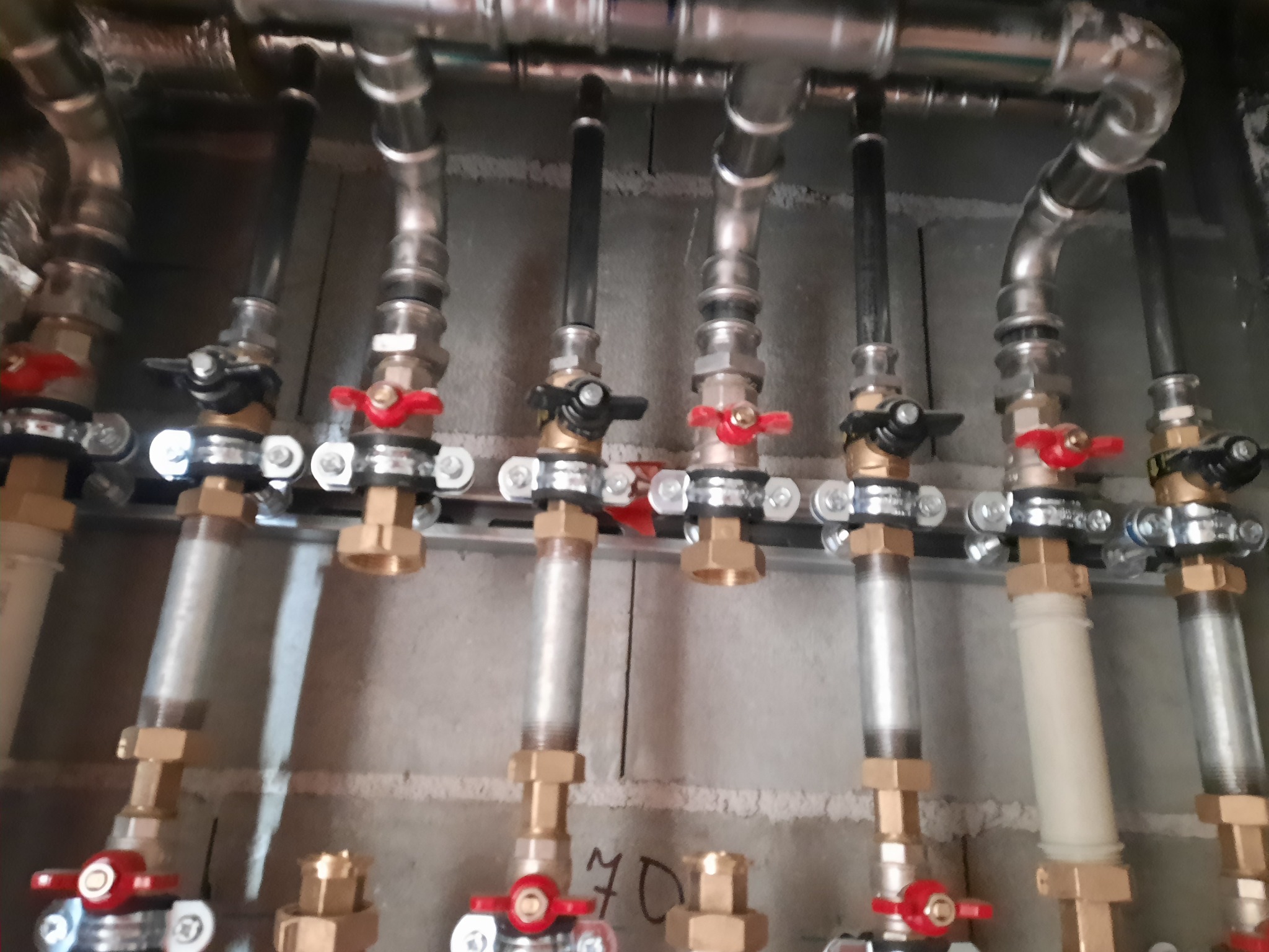 Rozdzielacz instalacji grzewczej z zaworami, rurami stalowymi i elementami z tworzywa sztucznego, zamontowany na szarej ścianie z betonu komórkowego.