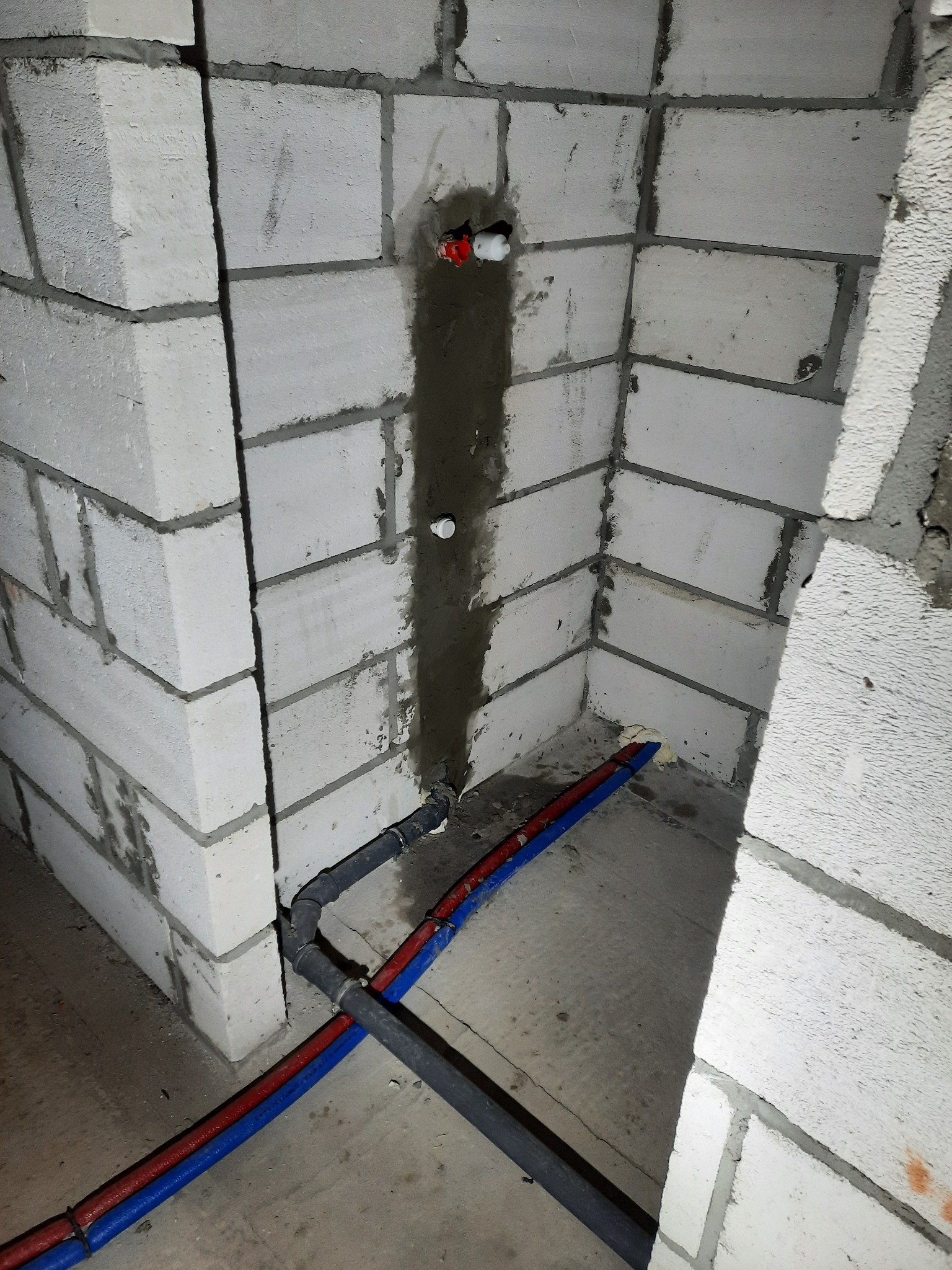 Surowa ściana z betonu komórkowego z bruzdą wypełnioną zaprawą, w której poprowadzone są rury instalacji wodnej i kanalizacyjnej z widocznymi złączkami i mocowaniami.