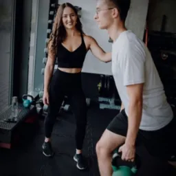 Trenerka personalna z długimi kręconymi włosami uśmiecha się, nadzorując ćwiczenie z kettlebell wykonywane przez mężczyznę w okularach w siłowni z rzędem hantli w tle.