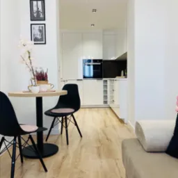 Jasne mieszkanie z aneksem kuchennym, okrągły stolik z czarnymi krzesłami, drewniana podłoga, białe ściany i czarno-białe zdjęcia na ścianie.