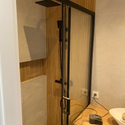 Creative Pro-Bud - Nowoczesna kabina prysznicowa z czarną armaturą i drewnianą okładziną ścienną, widoczny odpływ liniowy w podłodze i fragment szafki z podłączonym urządzeniem elektrycznym.