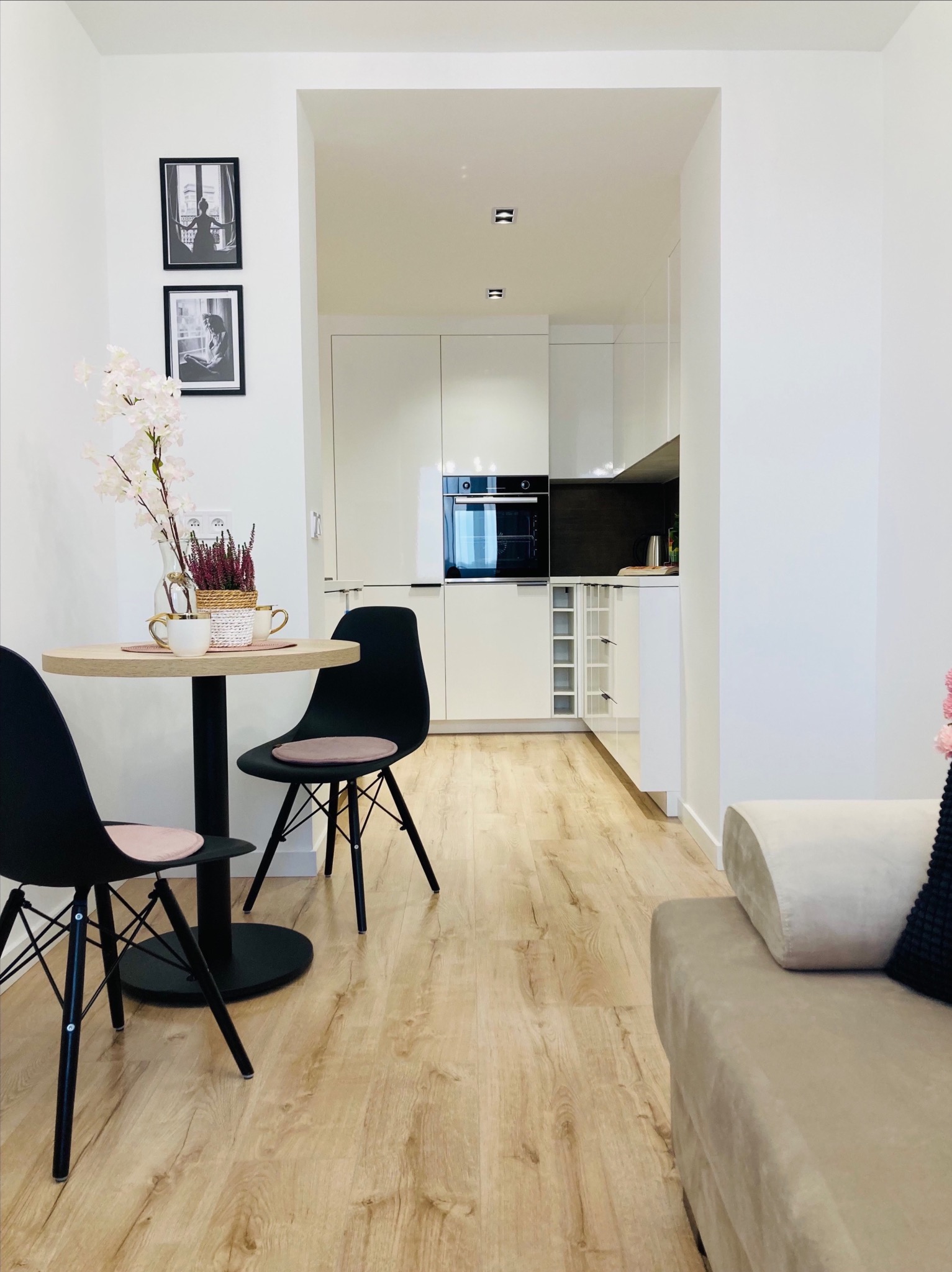 Jasne wnętrze mieszkania z drewnianą podłogą, okrągłym stolikiem z czarnymi krzesłami i widokiem na białą, nowoczesną kuchnię z zabudową.