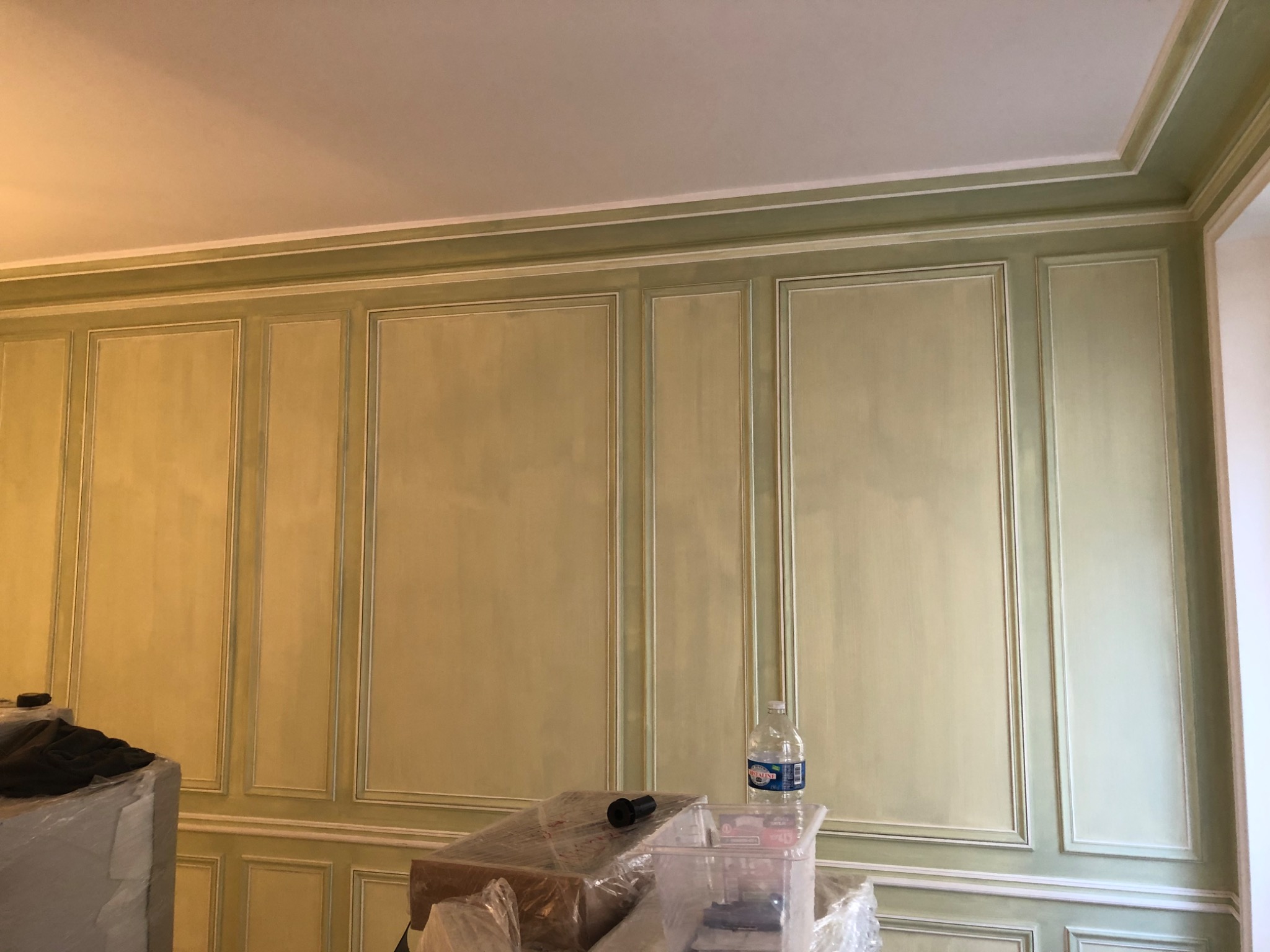 Ściana z dekoracyjnymi panelami w odcieniach zieleni i bieli, widoczna butelka wody mineralnej i materiały budowlane zakryte folią.