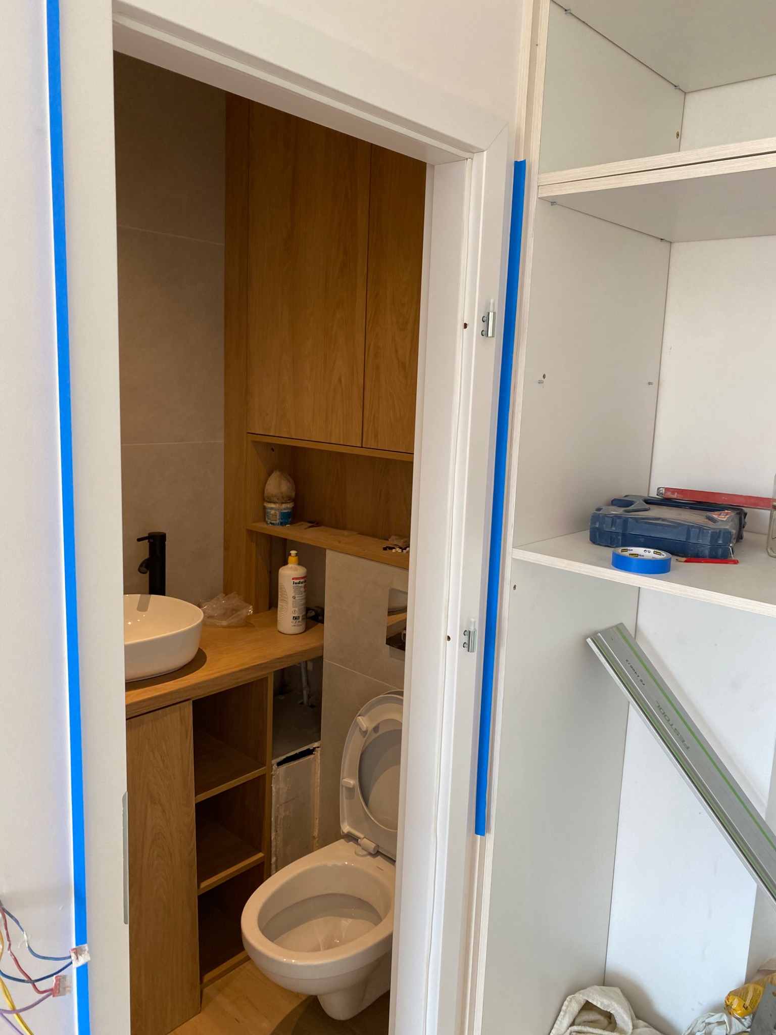 Wnętrze łazienki w trakcie prac wykończeniowych: widoczna umywalka z czarną armaturą, toaleta z otwartą klapą, zabudowa z drewnianymi szafkami i półkami oraz częściowo zamontowane białe regały...