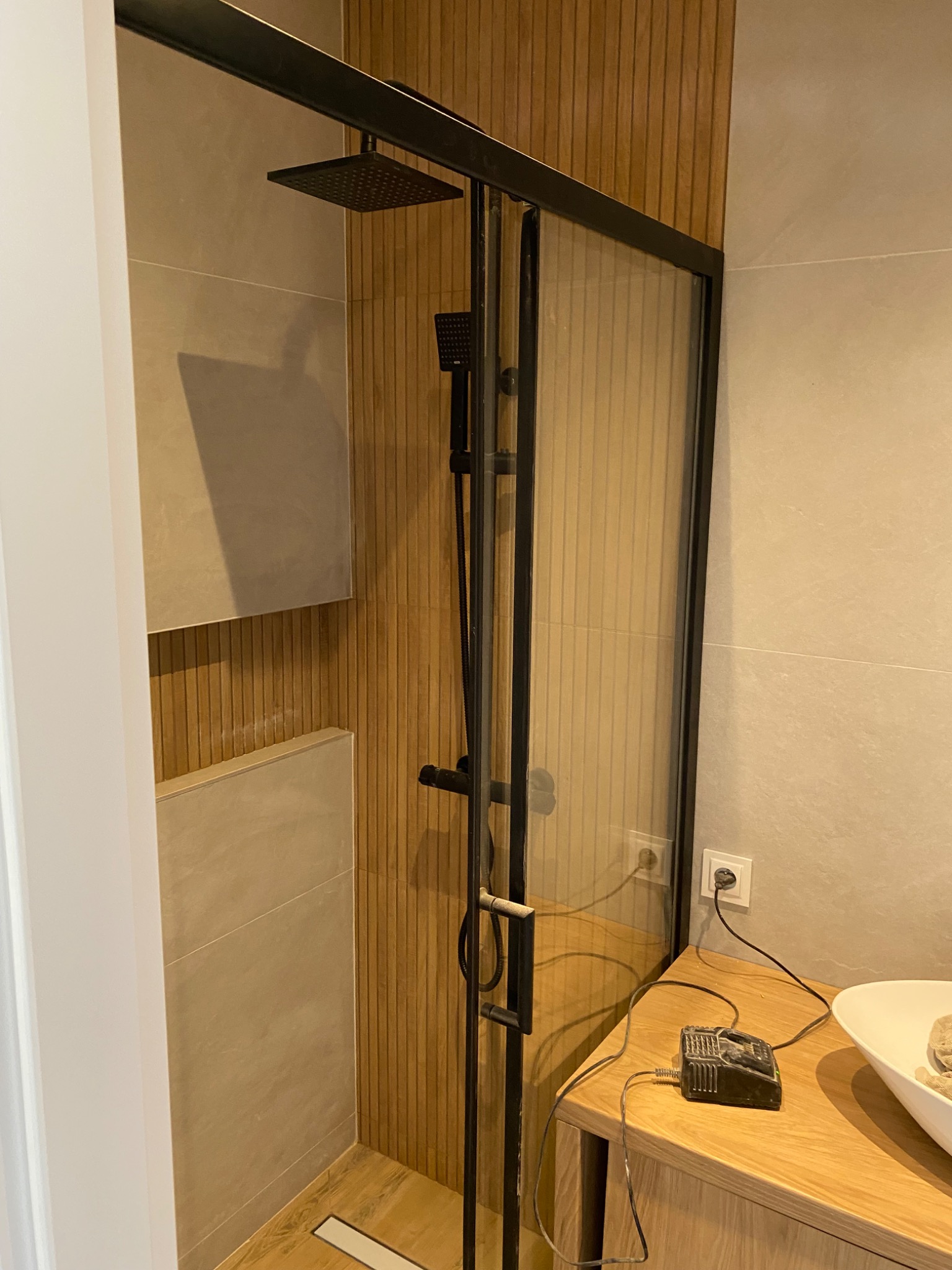 Nowoczesna kabina prysznicowa z czarną armaturą i drewnianą okładziną ścienną, widoczny odpływ liniowy w podłodze i fragment szafki z podłączonym urządzeniem elektrycznym.