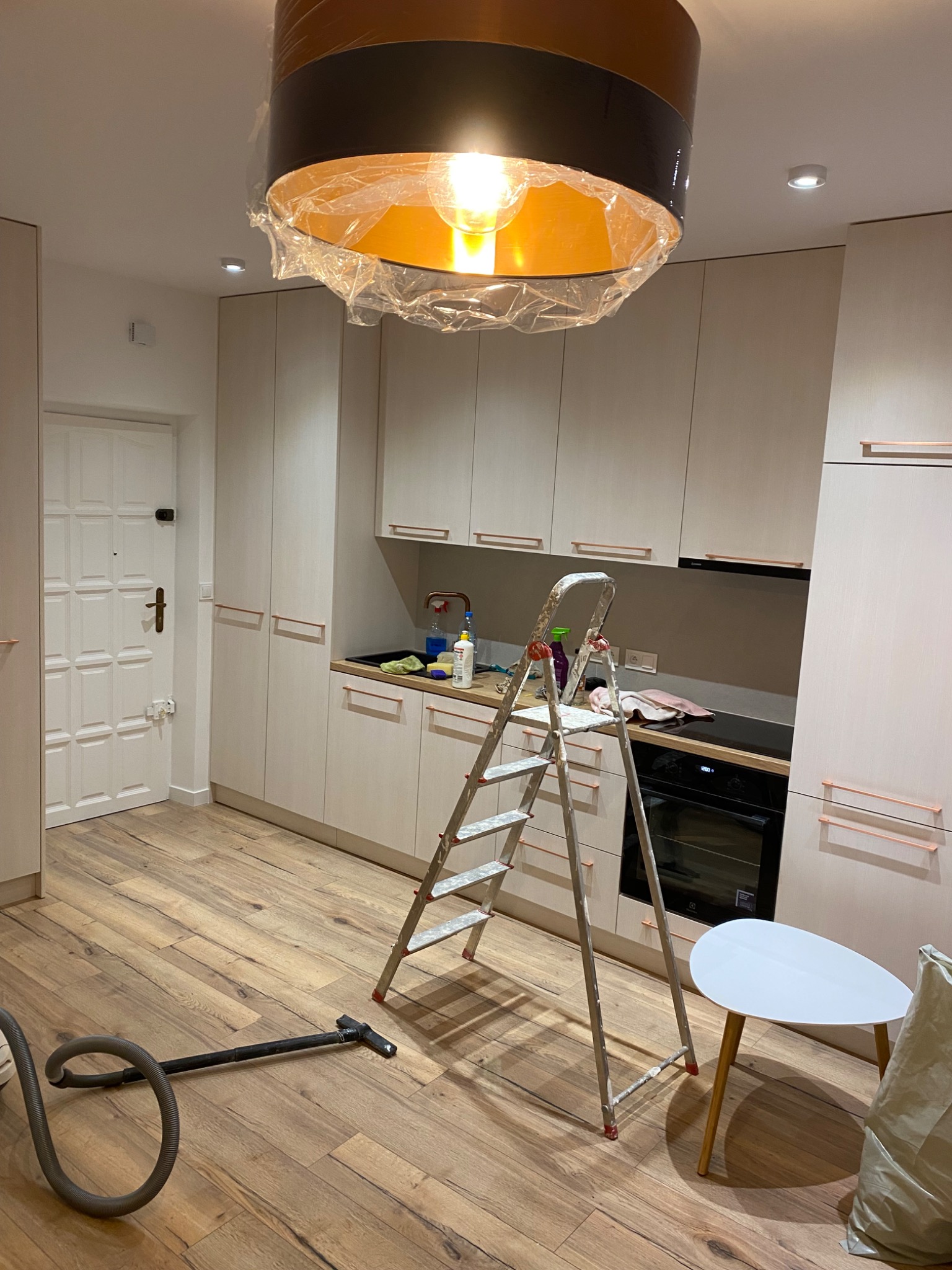 Kuchnia w trakcie remontu: drabina aluminiowa, odkurzacz na parkiecie, lampa z folią ochronną, meble kuchenne w jasnych odcieniach z miedzianymi uchwytami.