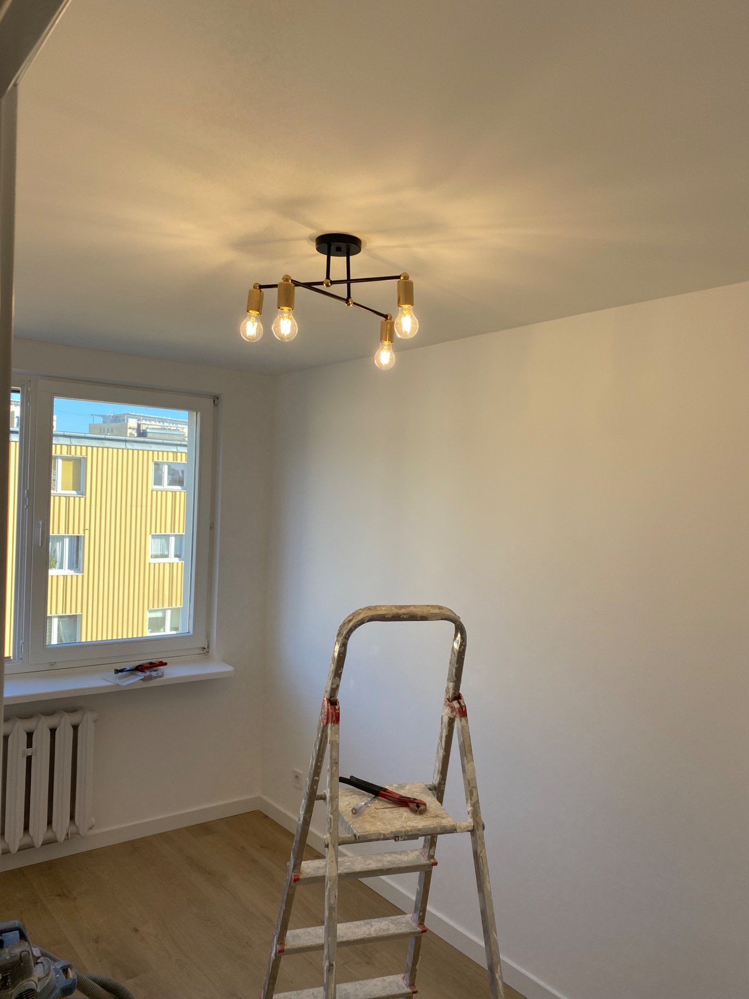 Wnętrze pokoju po malowaniu ścian, z nowoczesną lampą sufitową, drabina malarska w rogu i widok na żółty budynek za oknem.