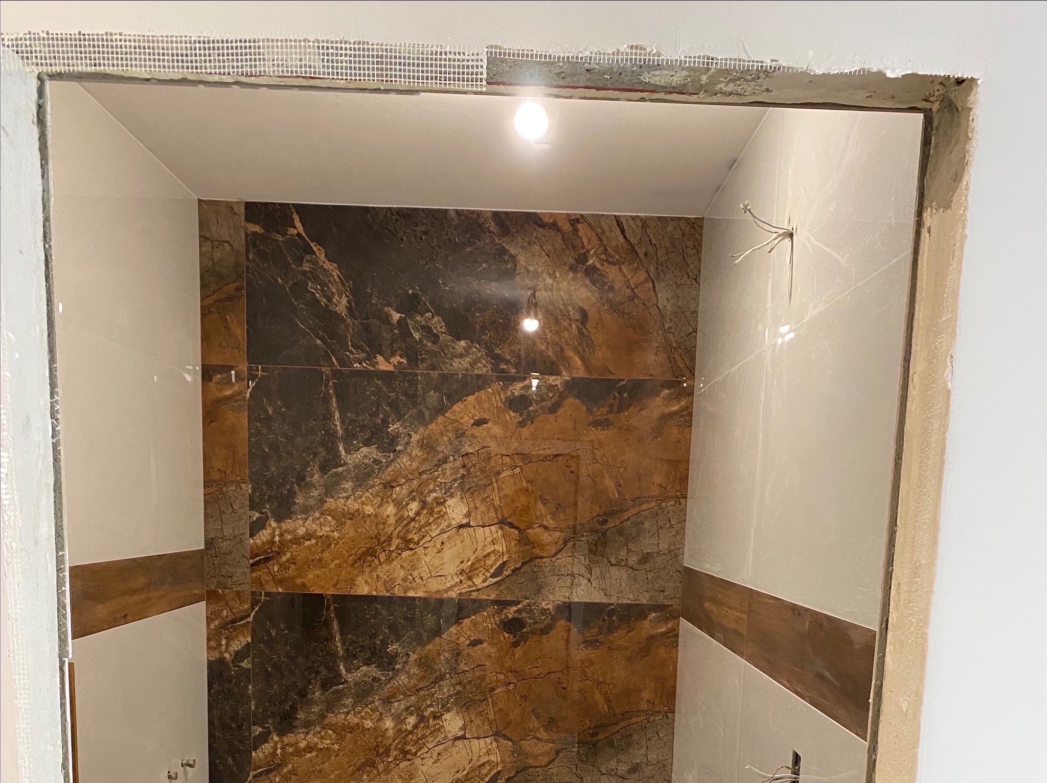 Wnętrze łazienki w trakcie wykańczania: ułożone płytki imitujące marmur i drewno, widoczne przewody elektryczne i siatka wzmacniająca na suficie.