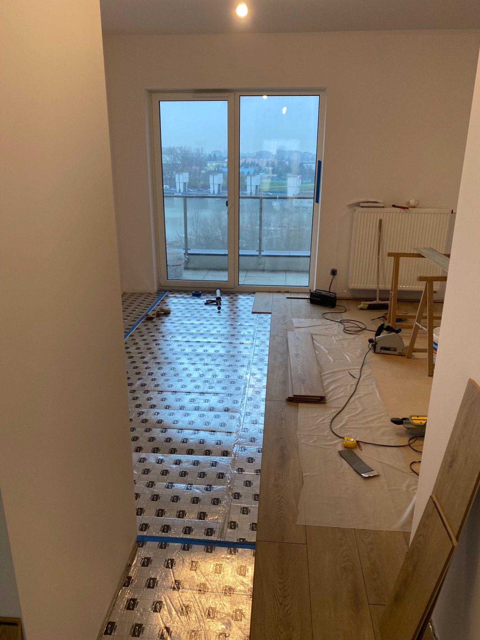 Przygotowanie do ułożenia paneli podłogowych w pokoju z balkonem: folia izolacyjna z taśmą, panele oraz narzędzia (piła, miarka).