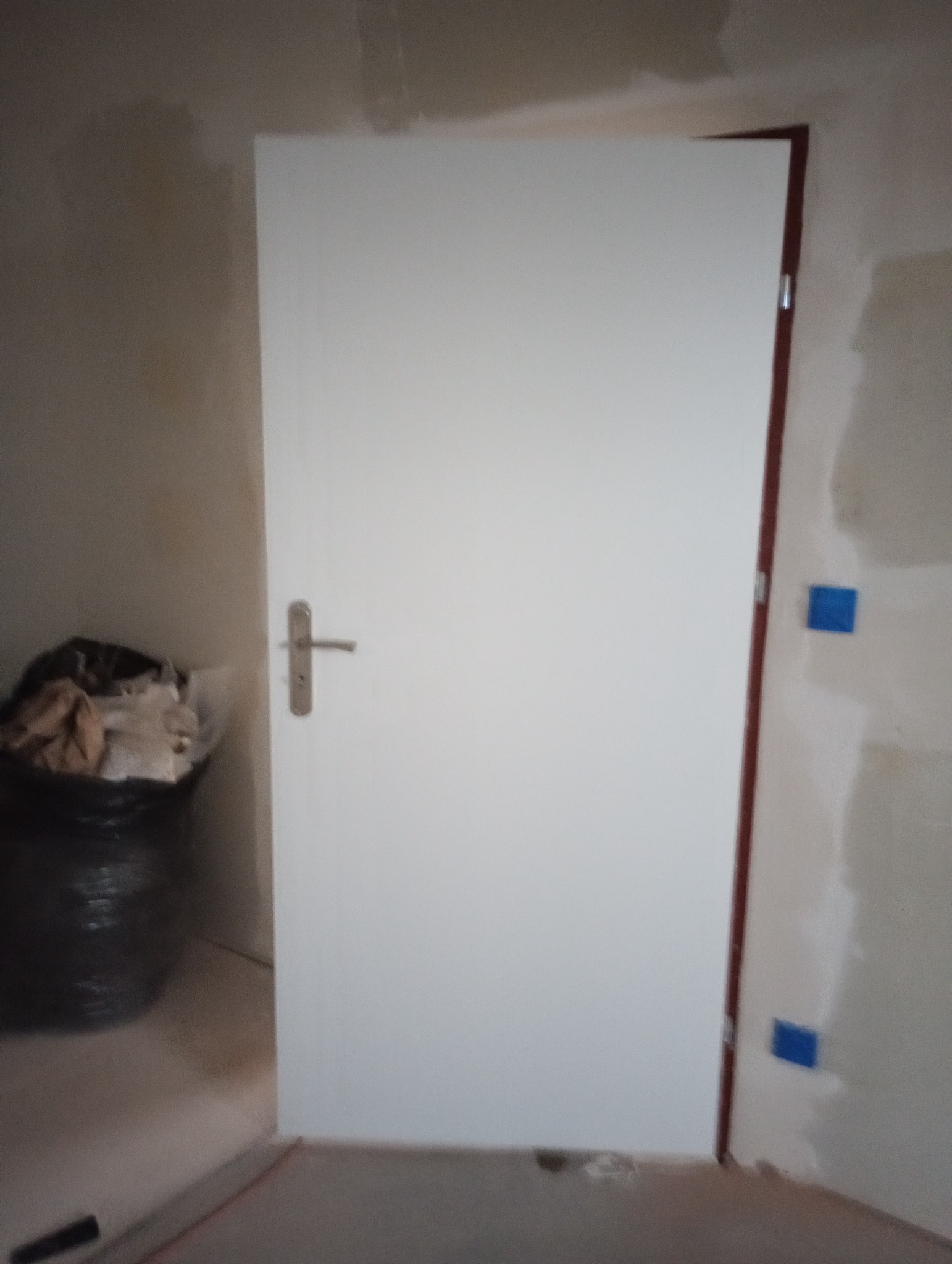 Białe drzwi wewnętrzne z klamką, zamontowane w surowym wnętrzu podczas remontu, zabezpieczone niebieską taśmą malarską na ścianie.