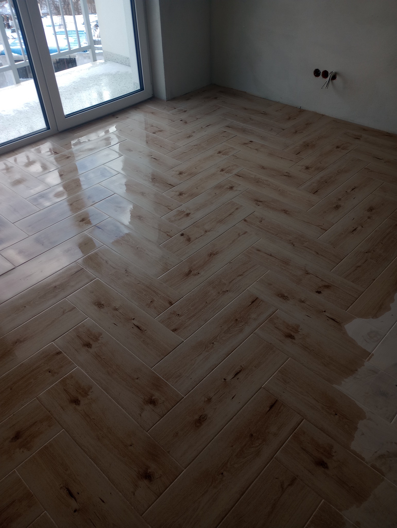 Ułożona, błyszcząca podłoga z płytek imitujących drewno w jodełkę, widoczne niezabezpieczone kable elektryczne w ścianie i okno balkonowe z widokiem na ośnieżony balkon.