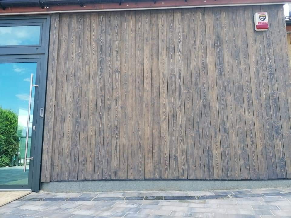 Drewniana elewacja budynku z pionowo ułożonych desek w odcieniach szarości i brązu, obok przeszklone drzwi wejściowe z metalowym uchwytem i system alarmowy na ścianie.