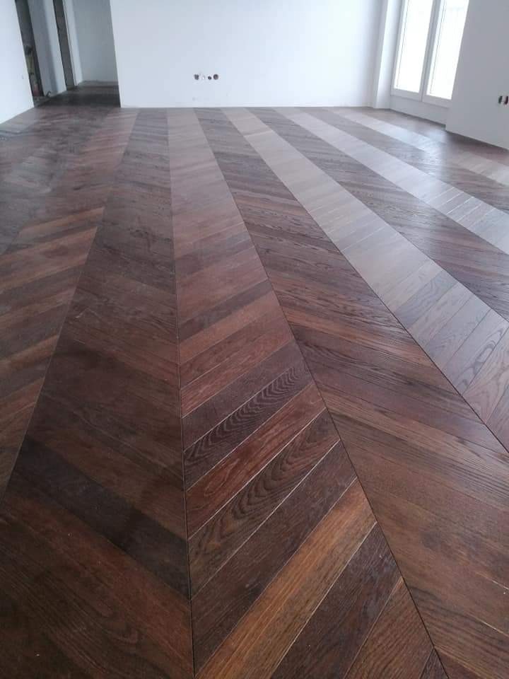 Ukośnie ułożony, ciemny parkiet w jodełkę na jasnym tle ściany z widocznymi gniazdkami elektrycznymi i oknem.