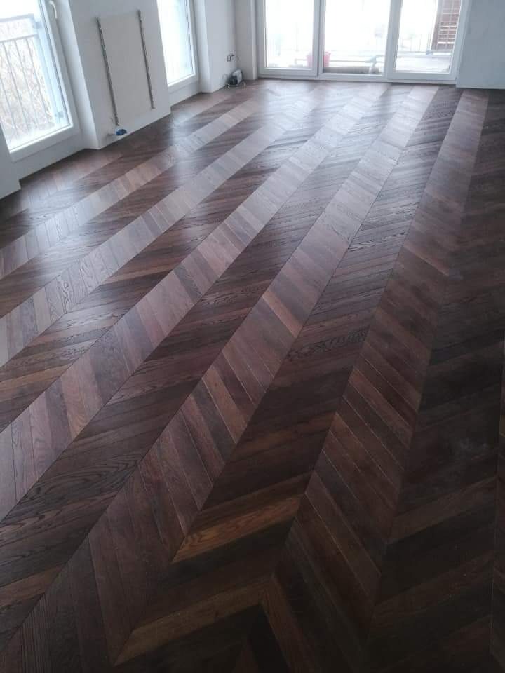 Ciemny parkiet ułożony we wzór jodełki w jasnym pomieszczeniu z dużymi oknami balkonowymi i grzejnikiem na ścianie.