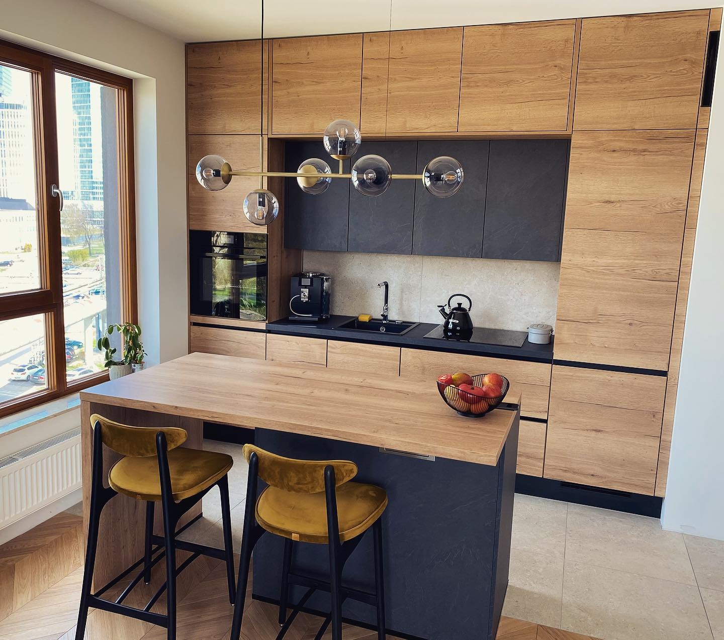 Nowoczesna kuchnia z drewnianymi frontami szafek, czarnymi blatami i wyspą z hokerami, oświetlona lampą z kulistymi kloszami, widok na miasto za oknem.